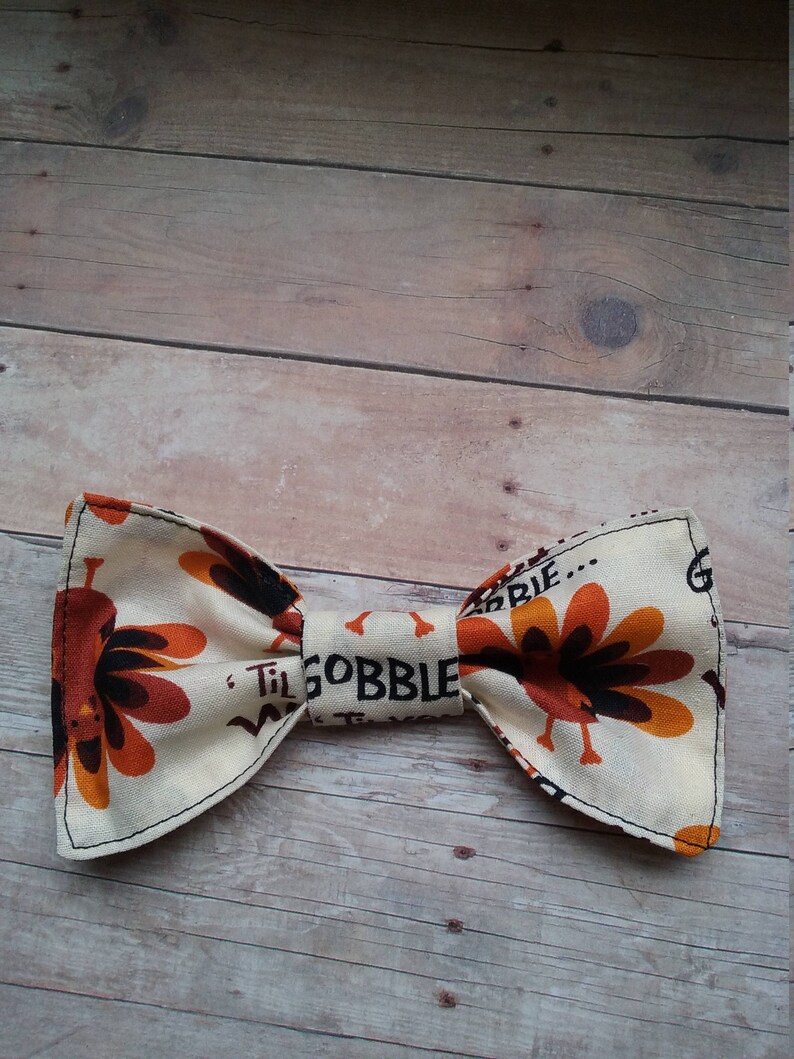 May include: A beige bow tie with a turkey print and the text "Til Gobble Til We Drop" in black.