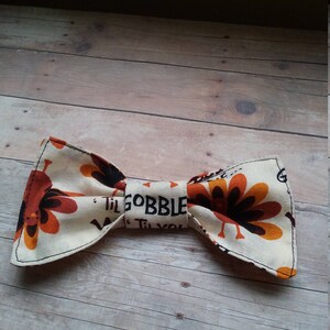 May include: A beige bow tie with a Thanksgiving theme. The bow tie features a pattern of turkeys with orange, brown, and black feathers. The text "Til Gobble Til You Wobble" is printed on the bow tie.