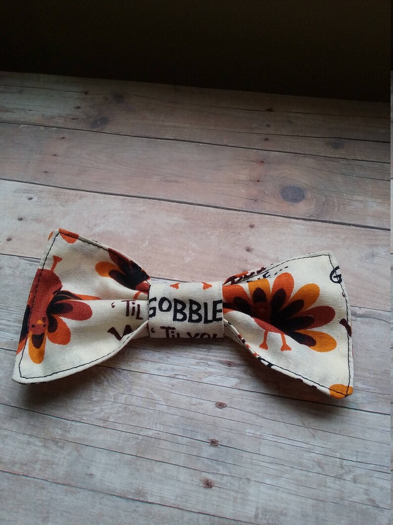 May include: A beige bow tie with a Thanksgiving theme. The bow tie features a pattern of turkeys with orange, brown, and black feathers. The text "Til Gobble Til You Wobble" is printed on the bow tie.