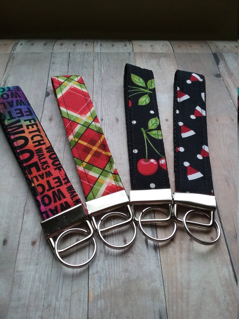 May include: Four colorful keychains with silver key rings. The keychains feature different patterns, including a red and green plaid, a black and white cherry design, a black and white Santa hat design, and a rainbow design with the words "FETCH" and "WALK" repeated.