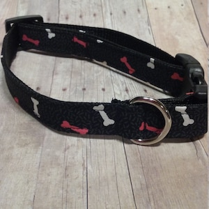 May include: Black dog collar with a white and red bone pattern. The collar has a silver metal D-ring.