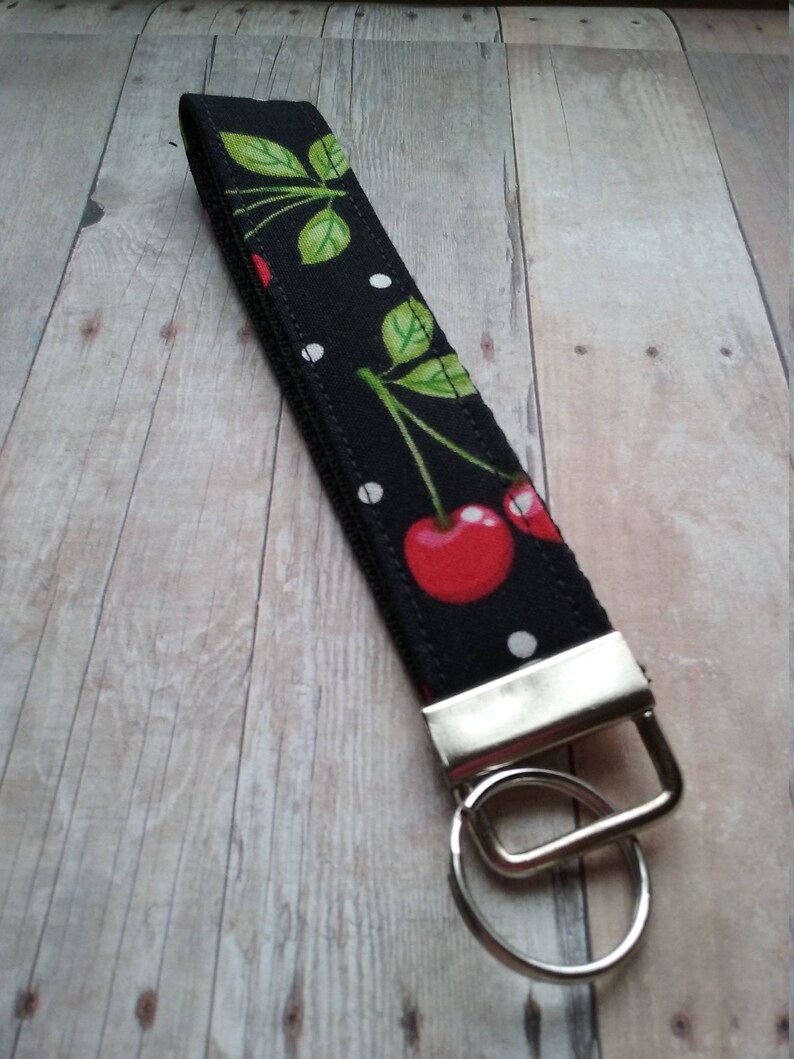May include: Black and white fabric key chain with a red cherry print and white polka dots. The key chain has a silver metal ring.