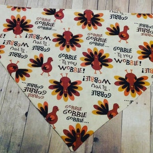 May include: A white bandana with a Thanksgiving turkey pattern. The bandana features a repeating pattern of turkeys with the text "Gobble Til You Wobble!" in brown and orange lettering.