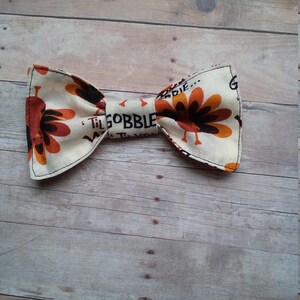 May include: A beige bow tie with a Thanksgiving turkey print. The bow tie features a brown turkey with orange feathers and the text "Til Gobble Til You Wobble".