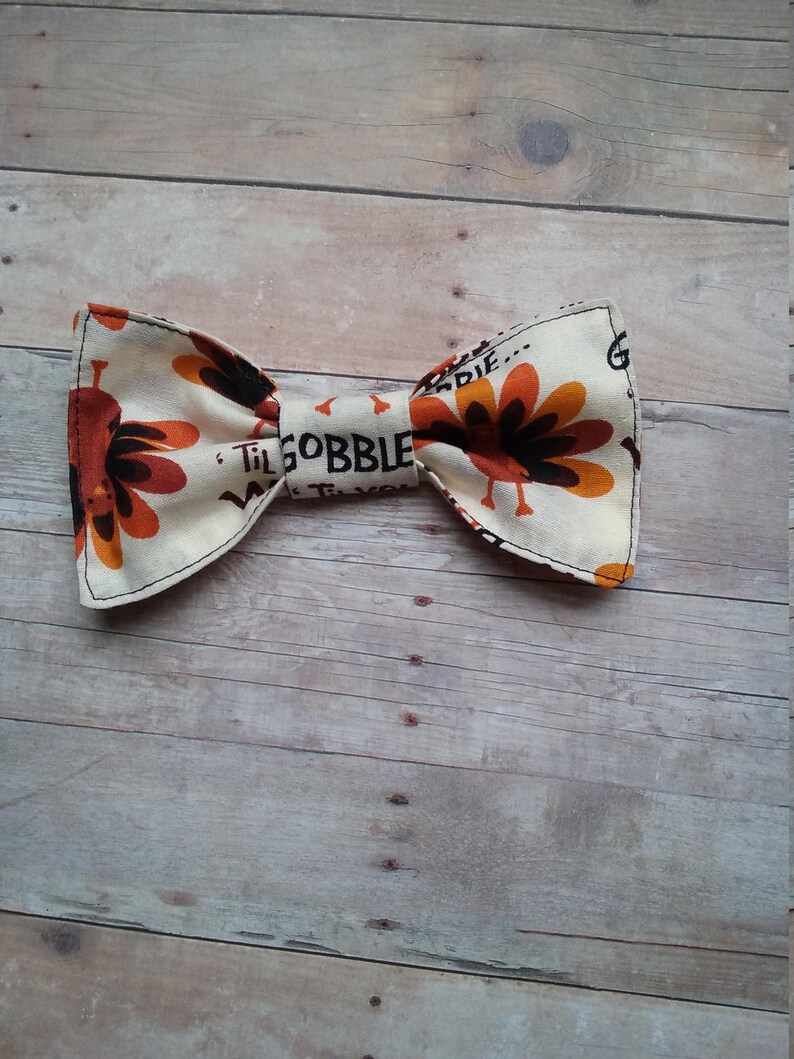 May include: A beige bow tie with a Thanksgiving turkey print. The bow tie features a brown turkey with orange feathers and the text "Til Gobble Til You Wobble".