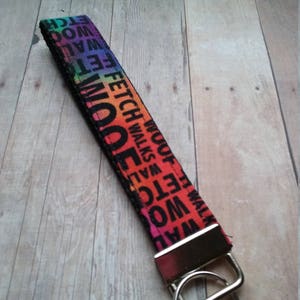 May include: A black key chain with a rainbow pattern and the words "FETCH WALK WOOF ETC" repeated. The key chain has a silver metal ring and a silver metal clip.