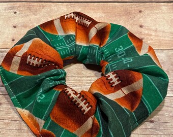 Puede incluir: Un scrunchie de pelo de tela verde y marrón con un patrón de fútbol. El scrunchie presenta un patrón repetido de balones de fútbol marrones sobre un campo verde.