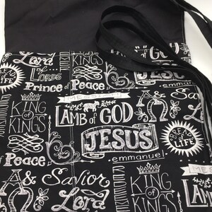 May include: Black fabric tote bag with white lettering. The lettering includes the words "Lord", "King", "Prince of Peace", "Lamb of God", "Jesus", "Savior", and "Emmanuel".