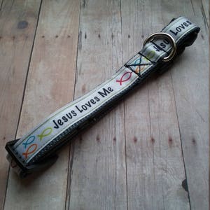 May include: White dog collar with black trim and the text "Jesus Loves Me" and colorful fish designs. The collar has a gold metal buckle and the text "Loves Me" on the buckle.