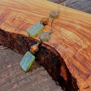 May include: A pair of handcrafted earrings featuring a combination of blue, orange, and green beads. The earrings have a floral design and are suspended from gold-colored ear wires. The beads have a textured appearance, adding to their unique charm.