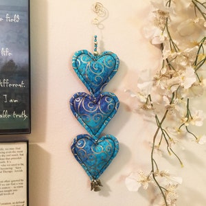 May include: A hanging decoration featuring three heart-shaped ornaments. Each heart is a vibrant teal color with gold swirling patterns. The hearts are connected and adorned with a small bell at the bottom. A decorative floral element is visible in the background.
