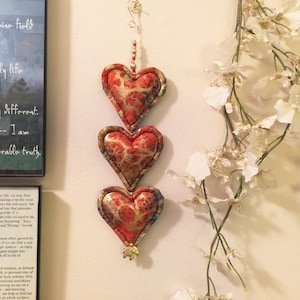 May include: Three heart-shaped ornaments with a red and gold patterned fabric, hanging vertically. Each heart is outlined with a dark stitch. A small gold charm hangs at the bottom. The background includes text and white floral accents.