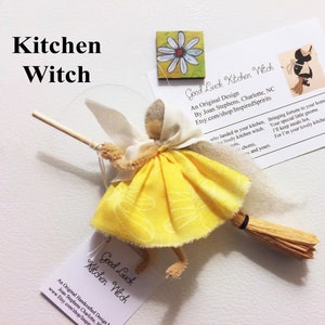 May include: A yellow kitchen witch doll with a brown broom and white wings. The doll is holding a wooden stick. The doll is on a white background with a business card that says "Good Luck Kitchen Witch" and has a website address.