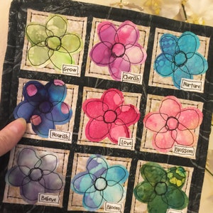May include: A black fabric square with nine embroidered flower patches. Each flower is a different color and has a word embroidered below it. The words are: Grow, Cherish, Nurture, Nourish, Love, Blossom, Believe, Bloom, and Plant.