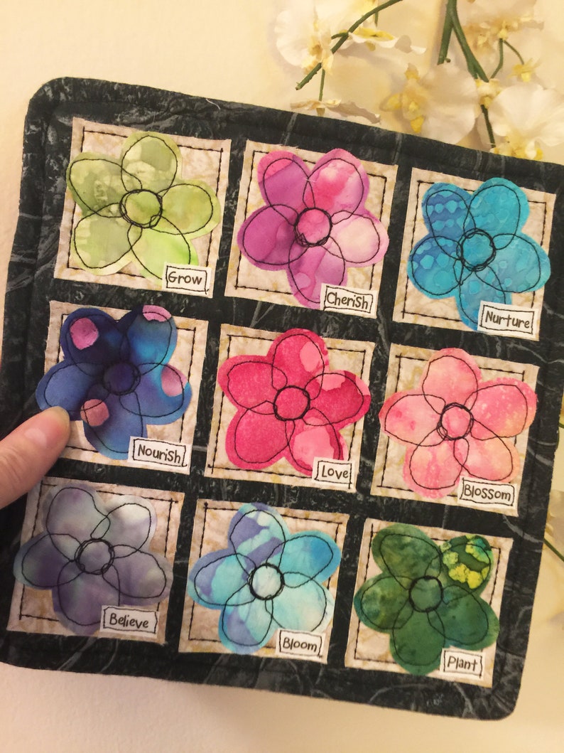 May include: A black fabric square with nine embroidered flower patches. Each flower is a different color and has a word embroidered below it. The words are: Grow, Cherish, Nurture, Nourish, Love, Blossom, Believe, Bloom, and Plant.