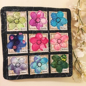 May include: A black fabric wall hanging with nine embroidered flowers, each with a different color and a word below it. The words are: Grow, Cherish, Nurture, Nourish, Love, Blossom, Believe, Bloom, and Plant.