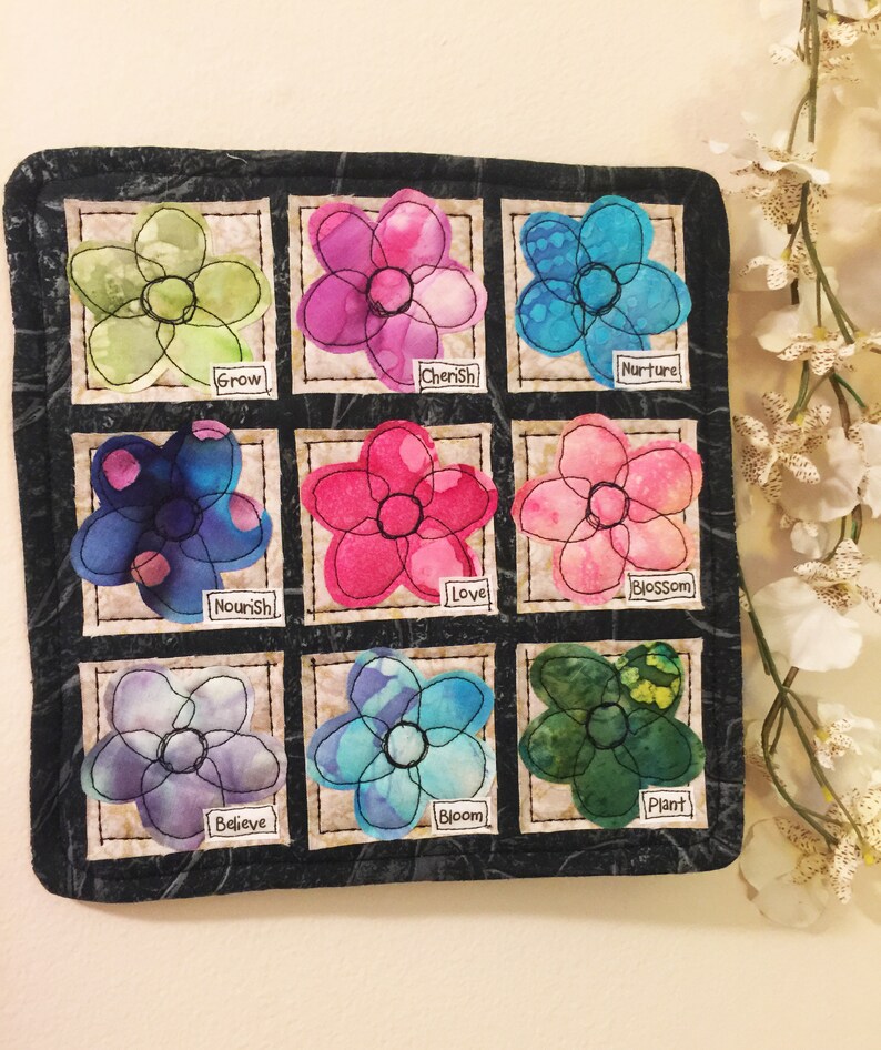 May include: A black fabric wall hanging with nine embroidered flowers, each with a different color and a word below it. The words are: Grow, Cherish, Nurture, Nourish, Love, Blossom, Believe, Bloom, and Plant.