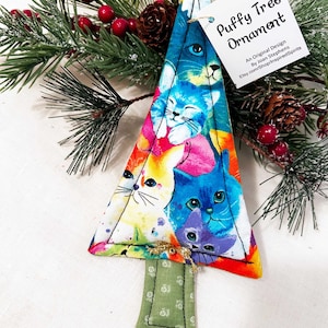 May include: A fabric Christmas tree ornament with a colourful print of cat faces in blue, pink, and yellow. The ornament has a green base and gold trim. The tag reads "Puffy Tree Ornament". The ornament is decorated with a sprig of pine and red berries.