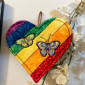 May include: A heart-shaped quilted fabric wall hanging featuring a rainbow-colored striped pattern with diagonal stitching. Two detailed butterfly appliques are centered on the surface, and a brown leather loop is attached to the top for hanging.