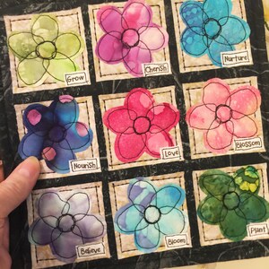 May include: A black fabric square with nine embroidered flower patches. Each flower is outlined in black thread and has a different color and pattern. Each patch has a word below it: Grow, Cherish, Nurture, Nourish, Love, Blossom, Believe, Bloom, Plant.