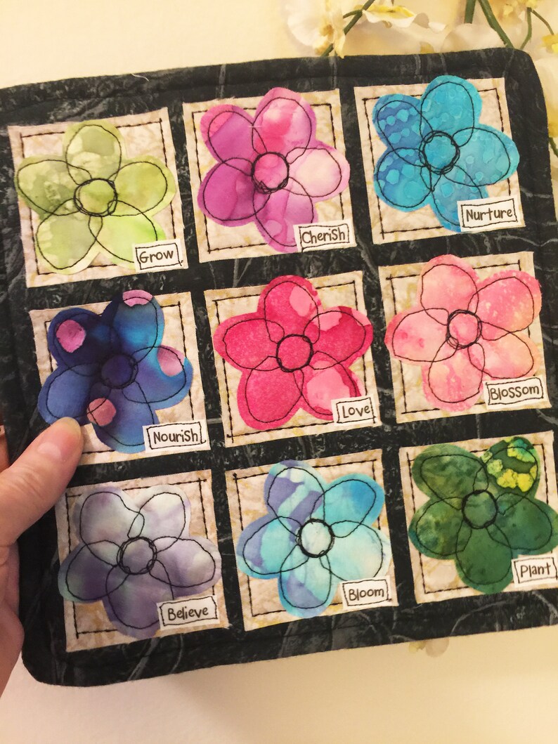 May include: A black fabric square with nine embroidered flower patches. Each flower is outlined in black thread and has a different color and pattern. Each patch has a word below it: Grow, Cherish, Nurture, Nourish, Love, Blossom, Believe, Bloom, Plant.