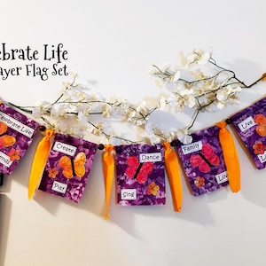 May include: A purple and orange prayer flag set with the words "Celebrate Life", "Friends", "Create", "Play", "Dance", "Sing", "Family", "Love", "Live", and "Laugh" printed on each flag. Each flag features a butterfly design.