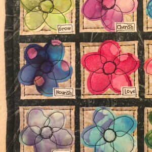 May include: A quilt with nine squares, each containing a different flower design. The flowers are all hand-stitched and have a watercolor look. The words "Grow", "Cherish", "Nourish", "Love", "Believe", "Bloom" are written on the quilt.