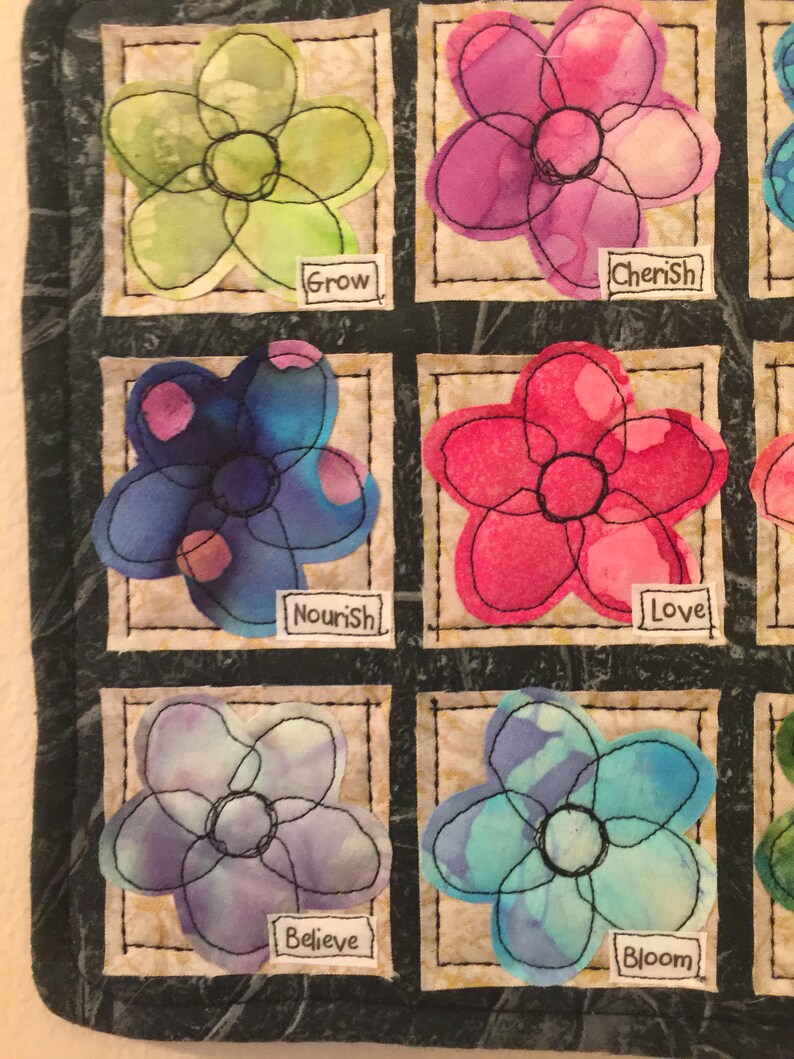 May include: A quilt with nine squares, each containing a different flower design. The flowers are all hand-stitched and have a watercolor look. The words "Grow", "Cherish", "Nourish", "Love", "Believe", "Bloom" are written on the quilt.