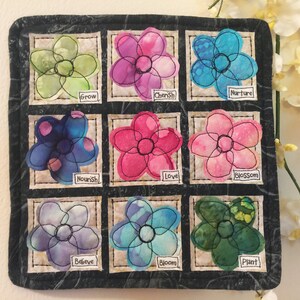 May include: A black fabric square with nine colorful fabric flowers, each with a different word embroidered on it. The words are: Grow, Cherish, Nurture, Nourish, Love, Blossom, Believe, Bloom, and Plant.