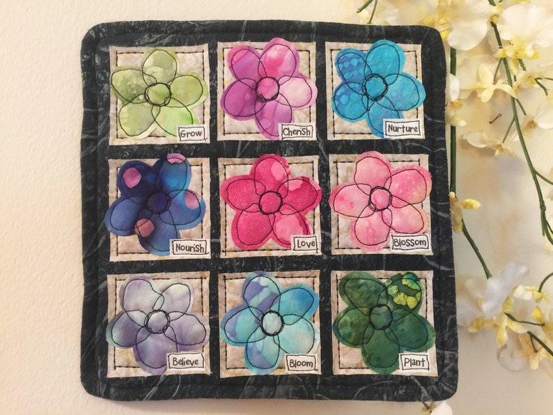 May include: A black fabric square with nine colorful fabric flowers, each with a different word embroidered on it. The words are: Grow, Cherish, Nurture, Nourish, Love, Blossom, Believe, Bloom, and Plant.