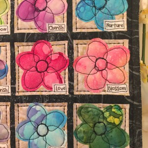 May include: A quilt block with nine squares, each containing a different flower design. The flowers are in various colors, including pink, blue, green, and purple. Each square has a white label with a word on it, such as "Cherish", "Nurture", "Love", "Bloom", and "Plant".