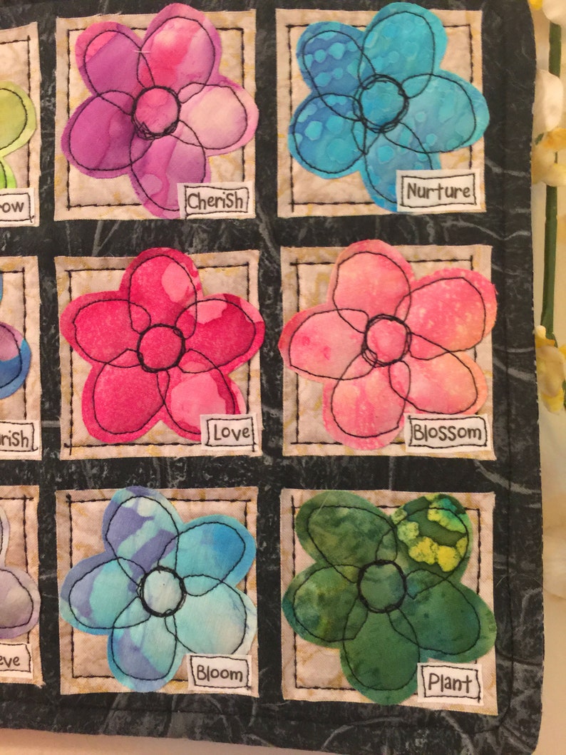 May include: A quilt block with nine squares, each containing a different flower design. The flowers are in various colors, including pink, blue, green, and purple. Each square has a white label with a word on it, such as "Cherish", "Nurture", "Love", "Bloom", and "Plant".