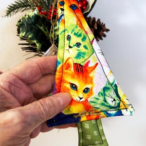 May include: A vibrant, triangular Christmas tree decoration with a cat-themed design. The ornament showcases a collage of cat faces in multiple colours. It has a green fabric base and a gold hanging string. The background includes foliage and red berries.
