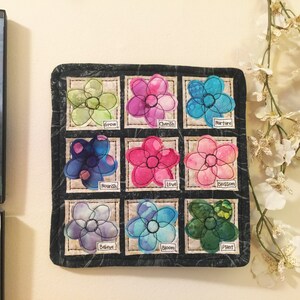 May include: A black fabric square with nine embroidered flower patches. Each flower is a different color and has a word underneath it, including "Grow", "Cherish", "Nurture", "Nourish", "Love", "Blossom", "Believe", "Bloom", and "Plant".