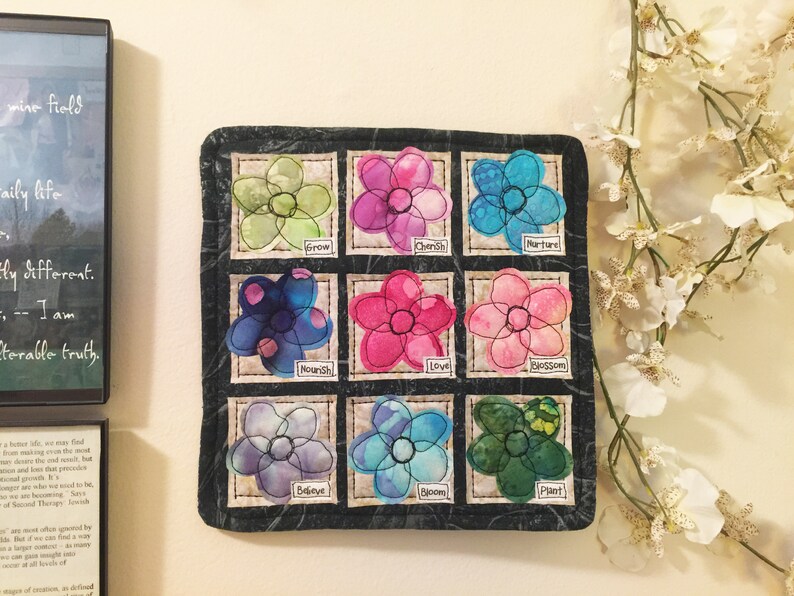 May include: A black fabric square with nine embroidered flower patches. Each flower is a different color and has a word underneath it, including "Grow", "Cherish", "Nurture", "Nourish", "Love", "Blossom", "Believe", "Bloom", and "Plant".