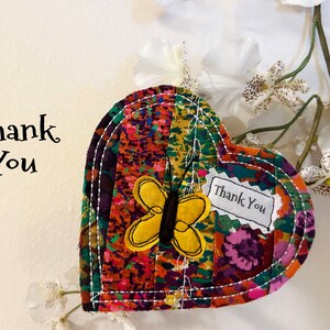 May include: A heart-shaped fabric ornament with a colorful patchwork design. A yellow butterfly with black accents is sewn onto the fabric. The ornament has a small tag that says "Thank You".