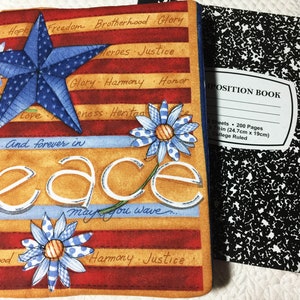 May include: A fabric notebook cover featuring a red, white, and blue star, blue and white daisies, and the text "...And forever in Peace may you wave". The cover displays horizontal stripes with words like "Brotherhood", "Glory", and "Justice". A black and white speckled composition book is visible next to it.