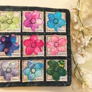 May include: A black fabric square with nine embroidered flower patches. Each patch has a different colored flower and a word underneath. The words are: Grow, Cherish, Nurture, Nourish, Love, Blossom, Believe, Bloom, Plant.