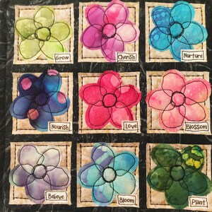 May include: A black fabric square with nine embroidered flower patches. Each flower is a different color and has a word underneath it. The words are: Grow, Cherish, Nurture, Nourish, Love, Blossom, Believe, Bloom, Plant.
