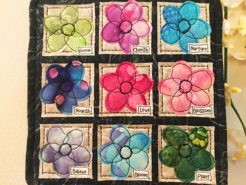 May include: A black fabric square with nine embroidered flower patches. Each flower is a different color and has a word underneath it. The words are: Grow, Cherish, Nurture, Nourish, Love, Blossom, Believe, Bloom, Plant.