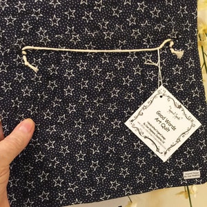 May include: Back view of a navy blue art quilt with a white star pattern. The quilt has a white string attached to it and a white tag with the text "Good Words Art Quilt" and "Handcrafted Original Design by Joan Stephens Ourto,  www.Etsy.com/shop/InspiredSpirits,  Gurnee, NC".