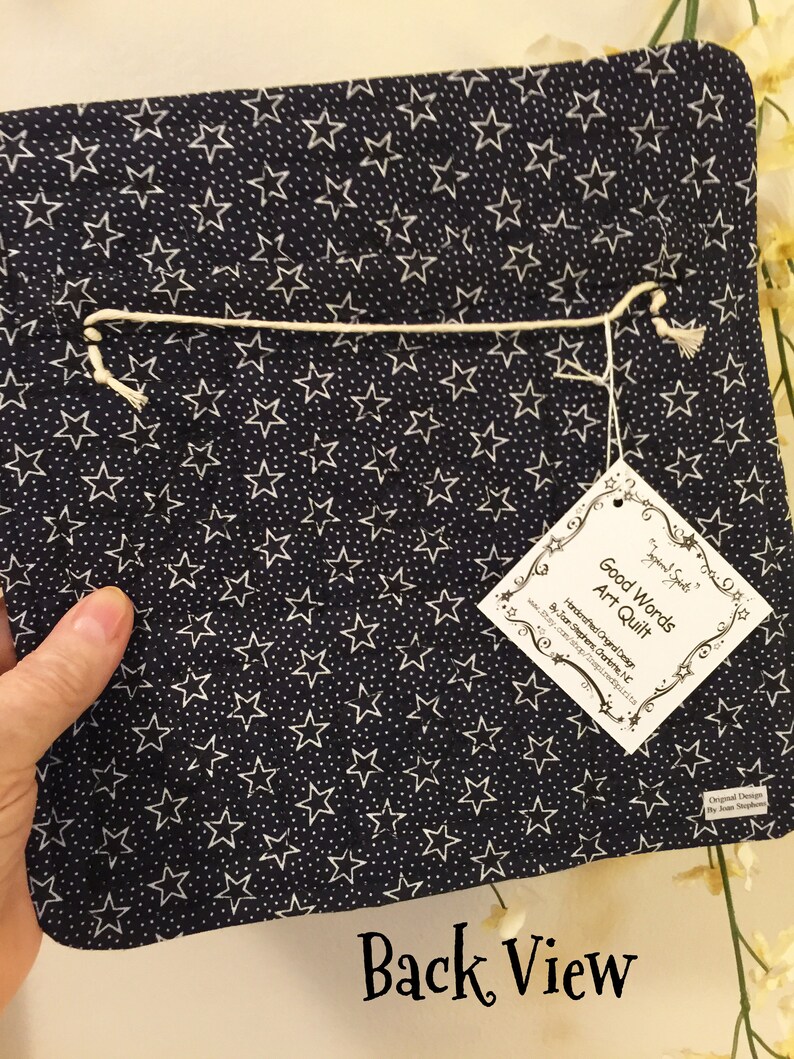 May include: Back view of a navy blue art quilt with a white star pattern. The quilt has a white string attached to it and a white tag with the text "Good Words Art Quilt" and "Handcrafted Original Design by Joan Stephens Ourto,  www.Etsy.com/shop/InspiredSpirits,  Gurnee, NC".