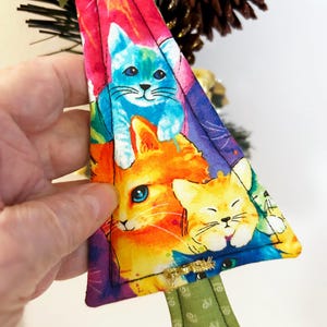 May include: A triangular Christmas tree decoration with a colourful print of cats in orange, blue, and yellow. The ornament has a green fabric base and gold trim. A festive item for cat lovers and holiday decorations.