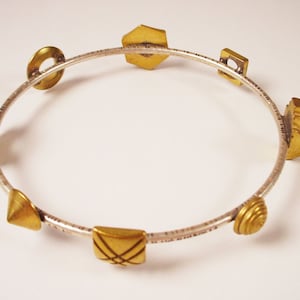 May include: A silver bangle bracelet with gold geometric shapes attached. The shapes include a hexagon, a circle, a square, a triangle, a crescent, and a spiral.