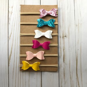 May include: A display of six colorful hair bows on elastic bands, attached to a brown cardboard backing. The bows are in shades of pink, blue, white, and yellow. The bows are made of glitter and textured materials.