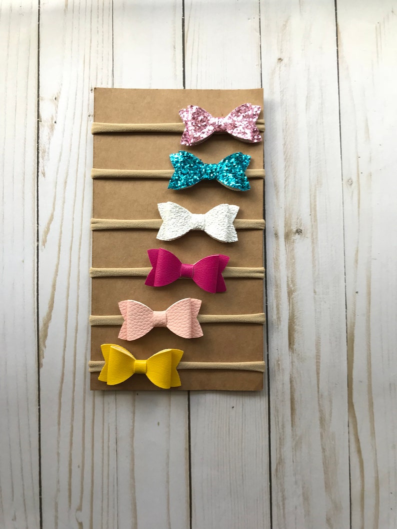 May include: A display of six colorful hair bows on elastic bands, attached to a brown cardboard backing. The bows are in shades of pink, blue, white, and yellow. The bows are made of glitter and textured materials.