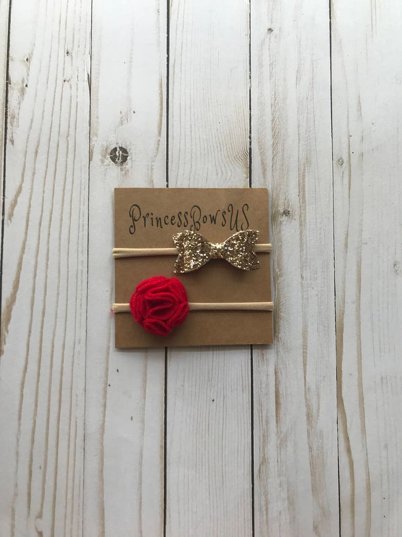 May include: Two baby headbands on a brown card. One headband has a gold glitter bow, and the other has a red felt flower. The card has the text "Princess Bows US" in gold script.