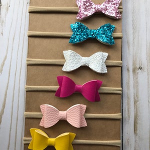 May include: A collection of six colorful baby headbands. The bows are in various colors, including pink glitter, turquoise glitter, white, fuchsia, light pink, and yellow. Each bow is attached to a neutral-colored elastic band and displayed on a brown cardboard backing.