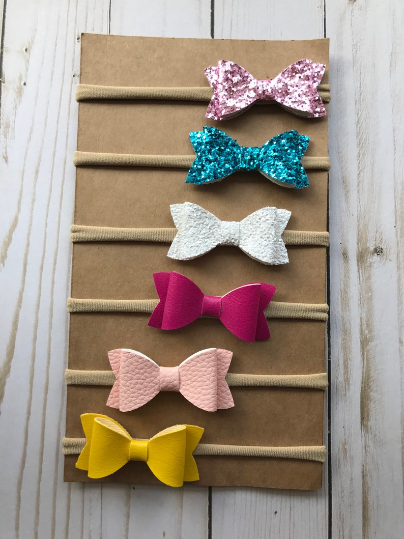 May include: A collection of six colorful baby headbands. The bows are in various colors, including pink glitter, turquoise glitter, white, fuchsia, light pink, and yellow. Each bow is attached to a neutral-colored elastic band and displayed on a brown cardboard backing.