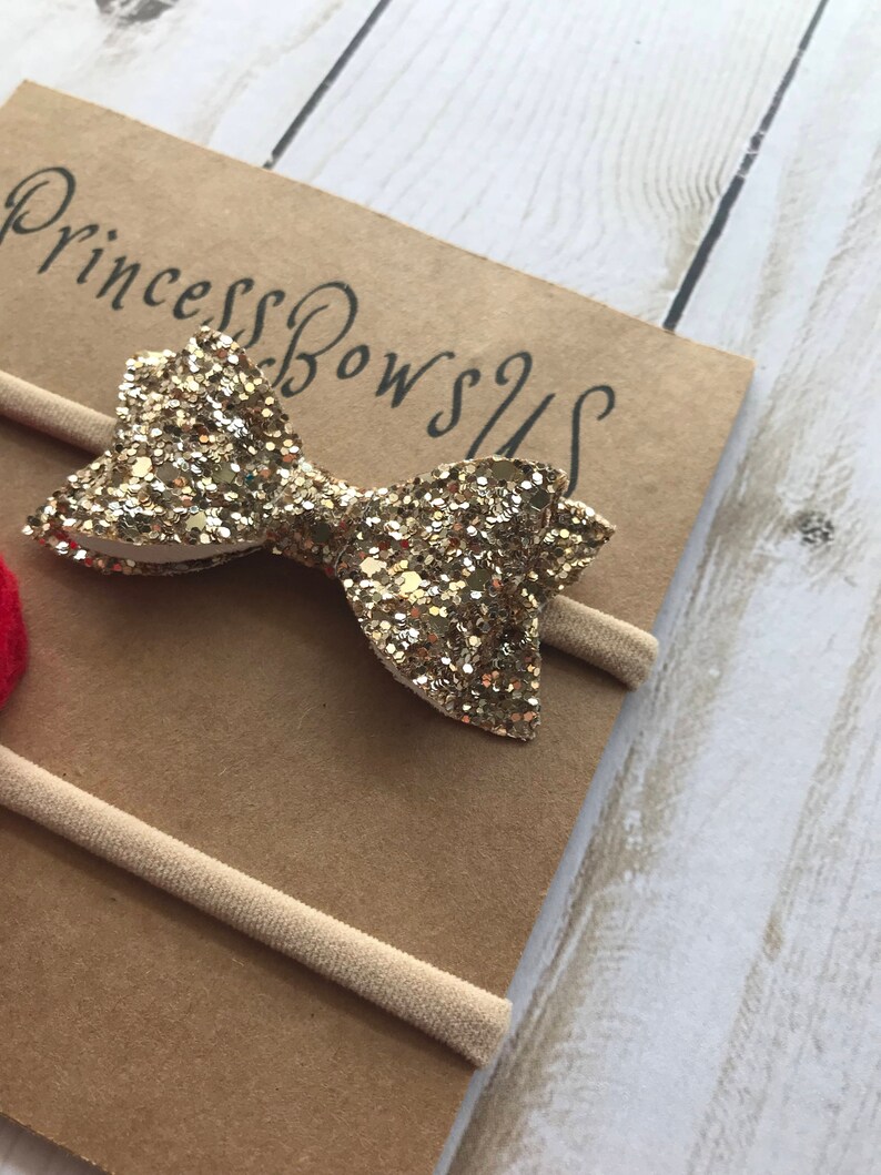 May include: A gold glitter bow headband with the text "Princess Bows US" on a brown card. The headband is made of a thin, beige elastic band.